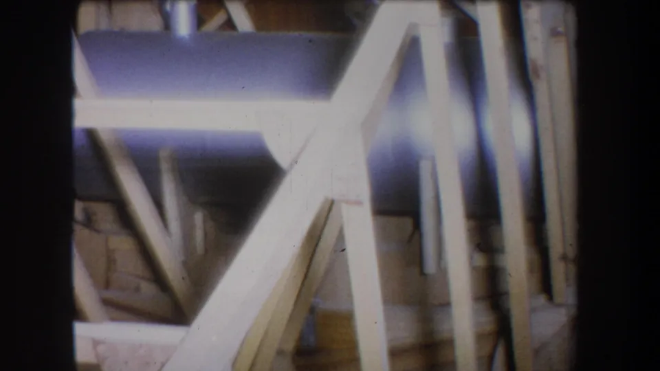 1965 California home-movie frame showing crossed wood supports, vertical framing pieces, and a bluish metal cylinder in an unfinished construction area.