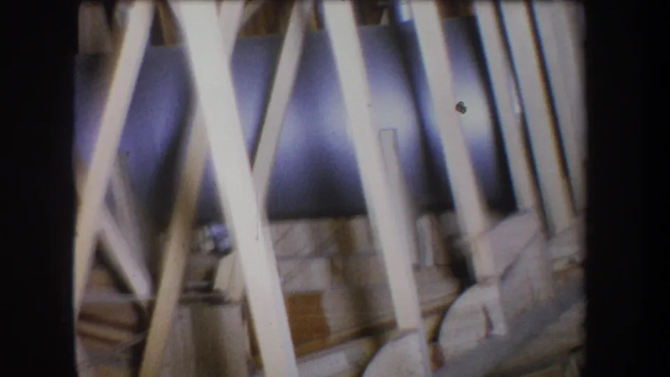Handheld 1965 8mm view of wooden structural braces with a thick dark pipe or cylindrical metal form in the background.