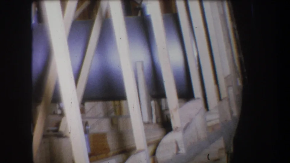 1965 home-movie frame with crossed pine beams, a silver pipe, and light wood planks viewed in soft focus.