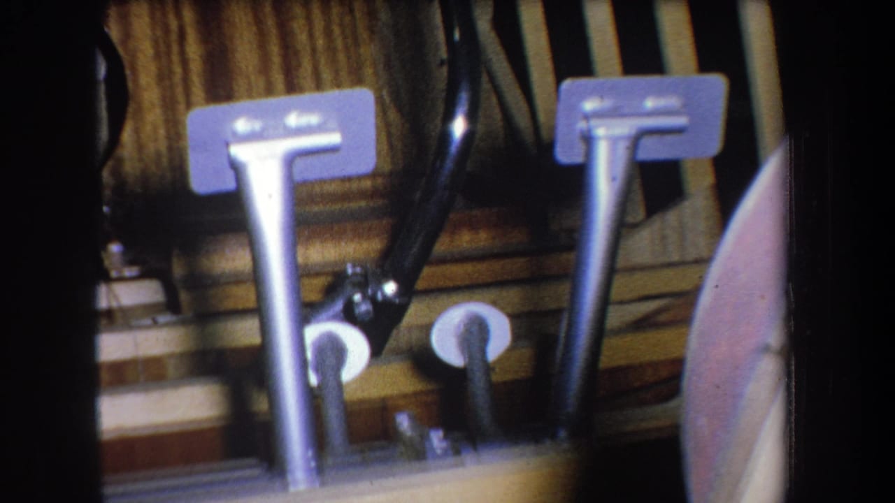1965 home-movie close-up of metal vehicle-style pedals, tubing, and linkage parts against a wood-toned panel in Yucaipa, California.