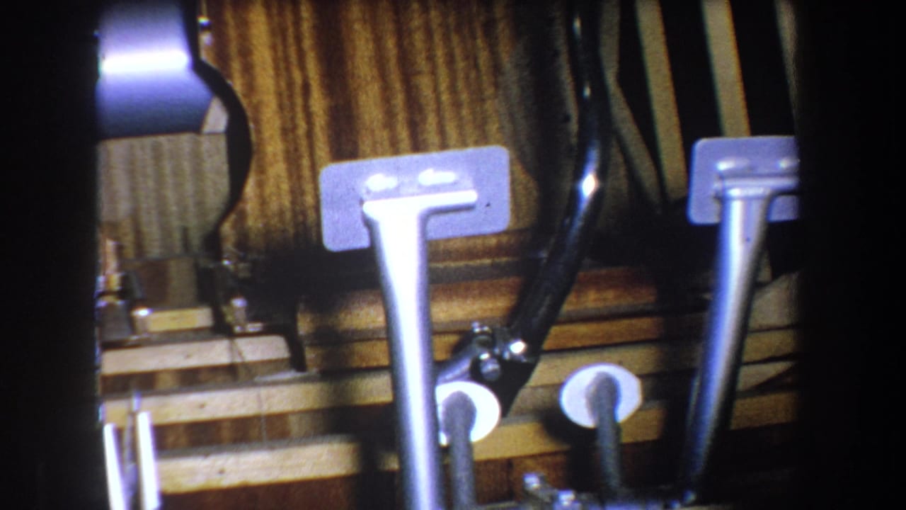 Close 8mm view of a vintage pedal assembly with metal foot controls, black tubing, and wood-grain backing from the 1965 reel.