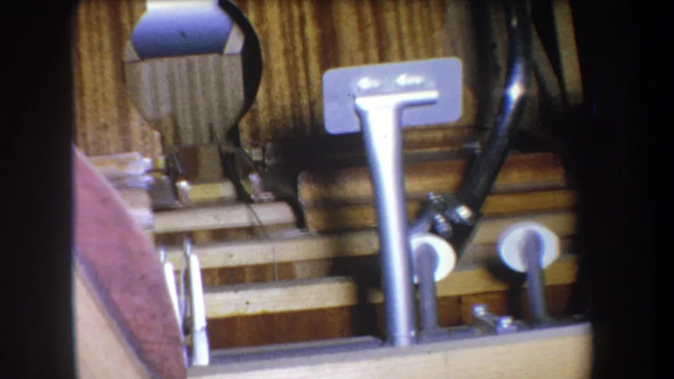 1965 8mm frame showing a metal pedal and exposed mechanical parts beside a pale pink curved surface and wood-toned backing.