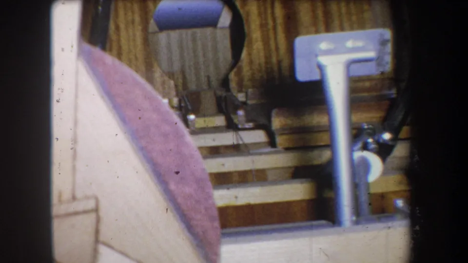 Close 1965 home-movie frame of a vintage mechanical pedal assembly with tubing, mounts, wood backing, and visible film wear.