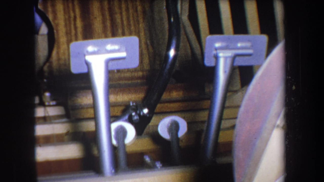 1965 Yucaipa 8mm frame showing vertical metal pipes, pedal-like levers, black tubing, and wood-paneled basement walls.