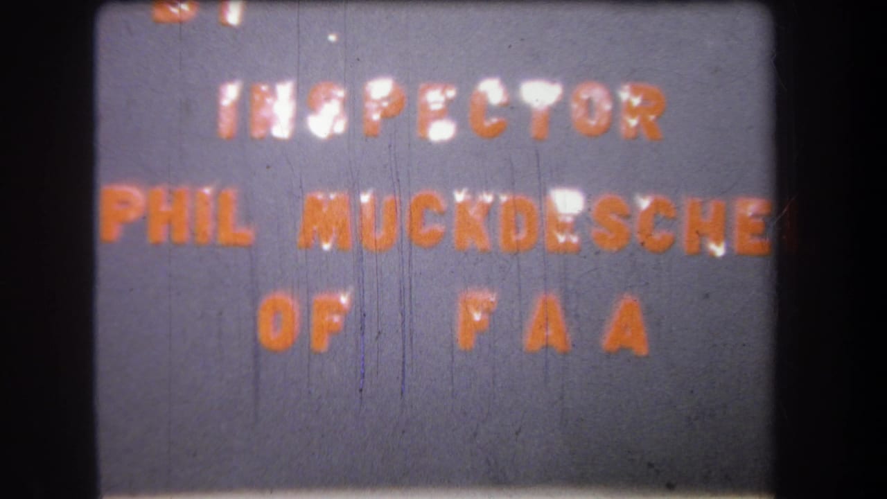 1965 8mm title card reading “BY INSPECTOR PHIL MUCKDESCH OF FAA” on a gray background, with soft focus and film wear.
