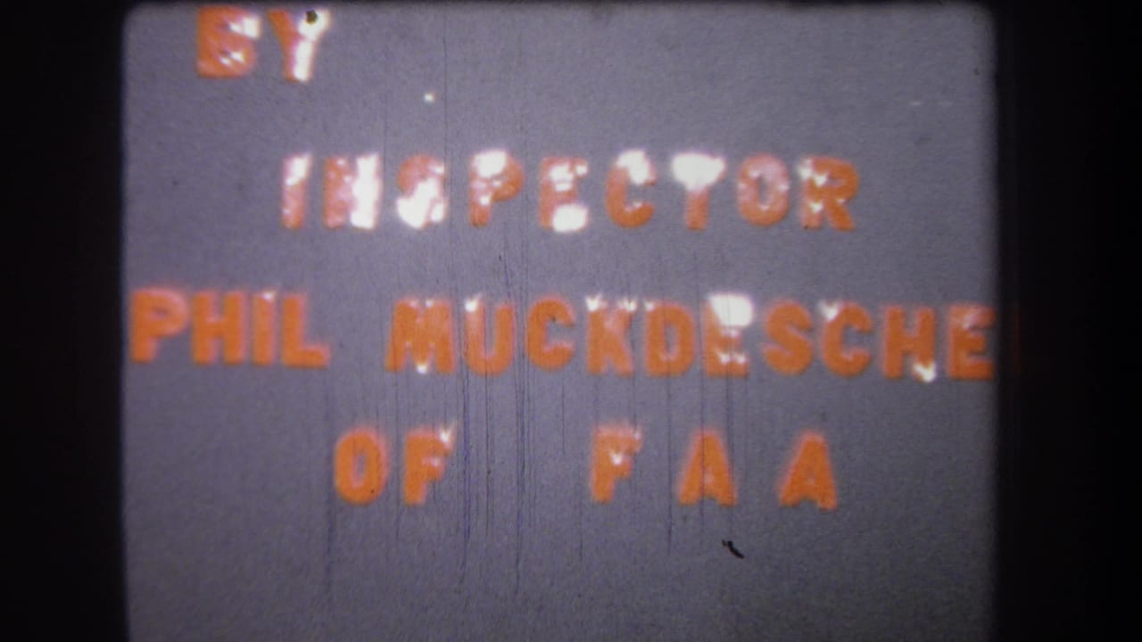 Mid-1960s 8mm frame showing the FAA inspector credit in orange letters on a gray card, bordered by dark film edges.
