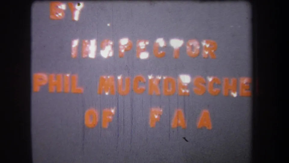 Mid-1960s 8mm frame showing the FAA inspector credit in orange letters on a gray card, bordered by dark film edges.