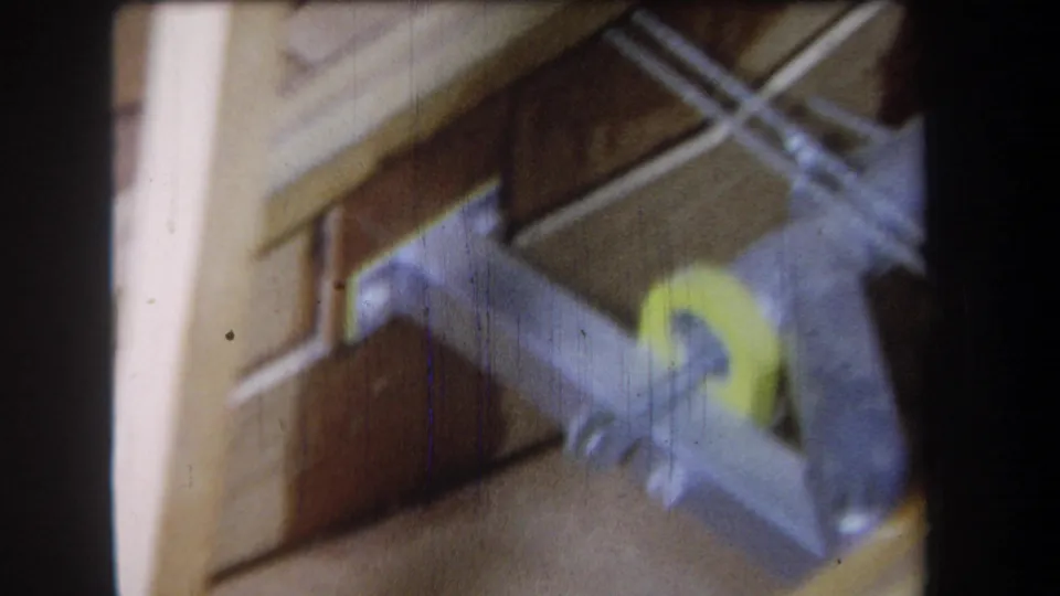 Close 1965 8mm frame of metal hardware or a tool against wood boards, with a yellow handle or grip visible.