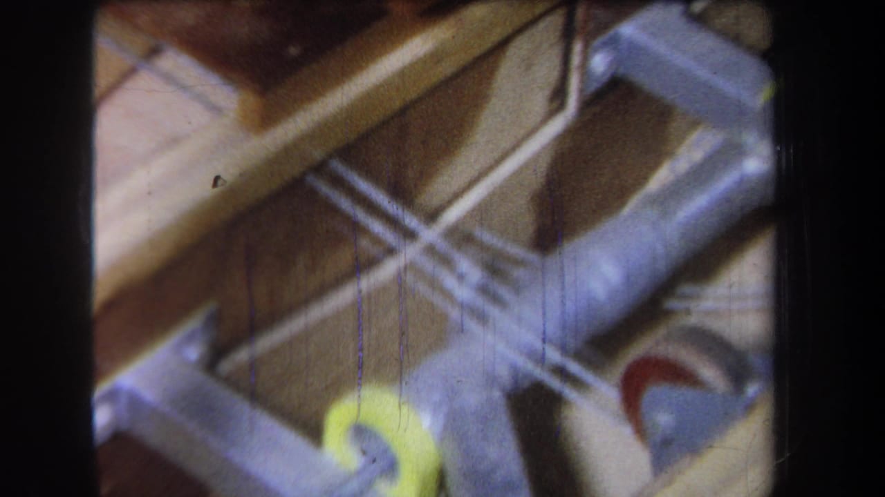 1965 home-movie frame from Volmer Yucaipa showing metal ducting, wood framing, and a yellow wheel-like component in an attic space.