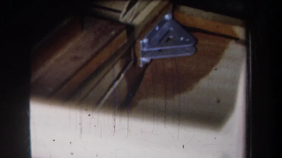 Final 1965 frame from the attic clip showing exposed wood, a metal bracket, pale surface material, and dark home-movie frame edges.