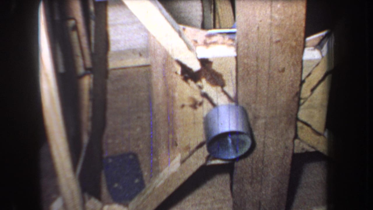 Close 1965 home-movie view of worn door wood, diagonal framing, a metal cylinder-shaped fixture, cracks, stains, and dim interior shadow.