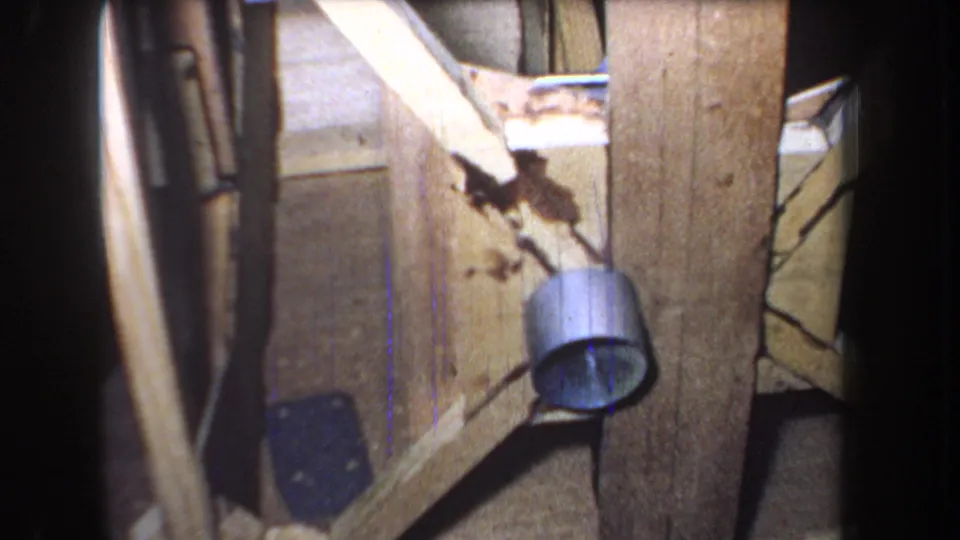 Close 1965 home-movie view of worn door wood, diagonal framing, a metal cylinder-shaped fixture, cracks, stains, and dim interior shadow.