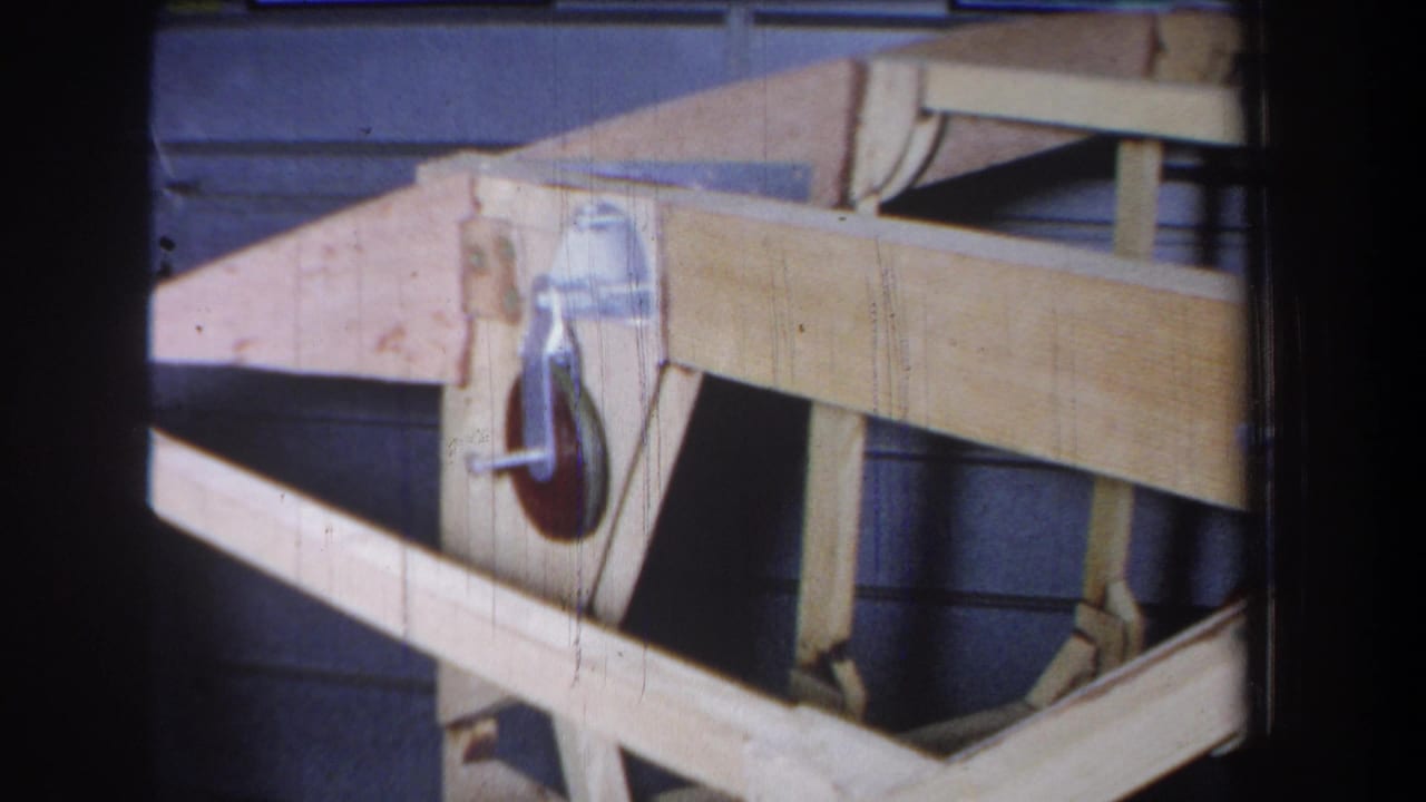Wooden angled structure with a metal pulley wheel mounted on a side bracket against blue-gray siding.