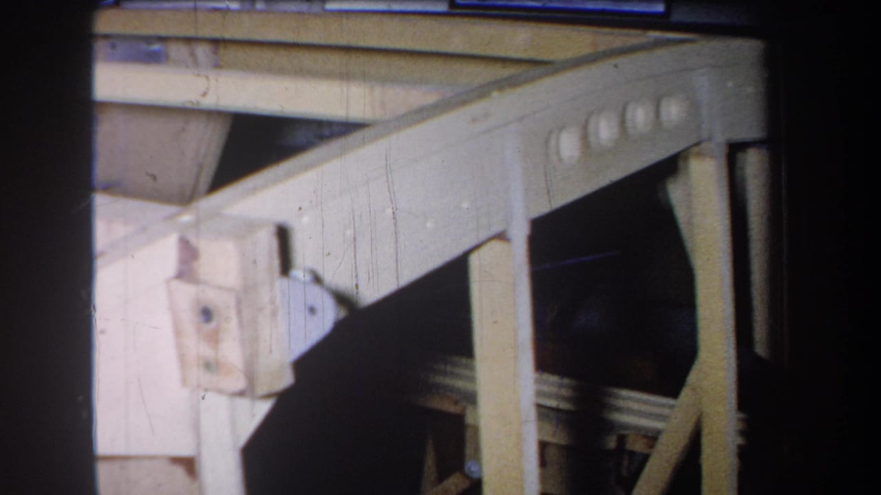 1965 Yucaipa home-movie frame showing a close section of wooden mechanical framing with metal hinge plates and dark gaps behind it.