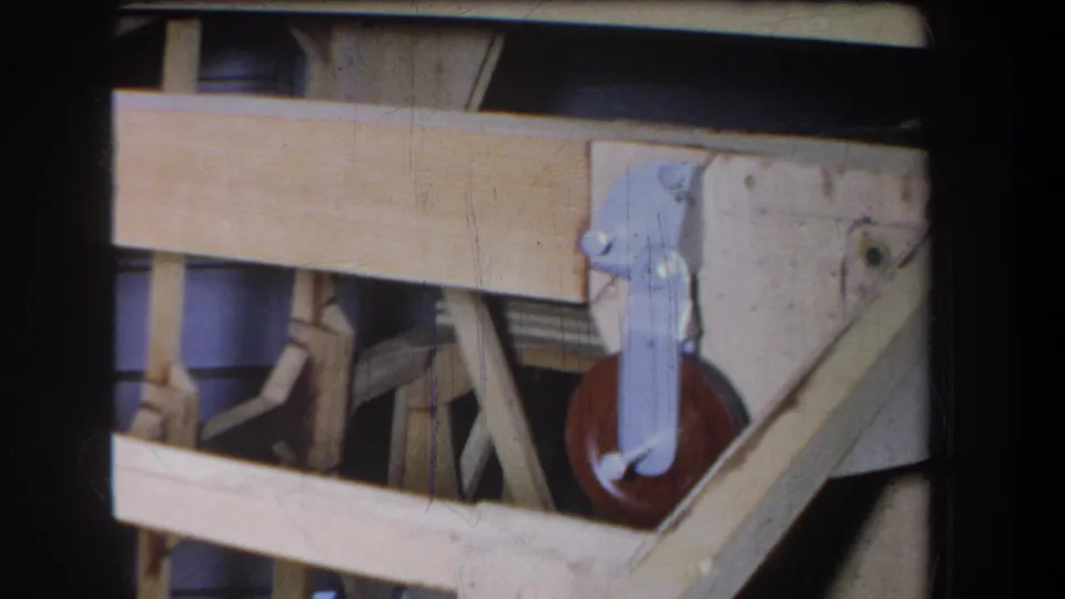 Close 1965 Yucaipa frame showing the mechanism's wooden rails, metal hinge hardware, and a red-brown wheel mounted near the lower right.