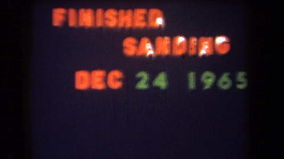 Projected 1965 home-movie title card with orange project text and green date against a dark, grainy field.