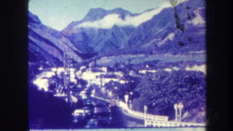 Elevated 1967 Hawaii view of cars on a valley road below mountains and low clouds, with buildings and vegetation visible.
