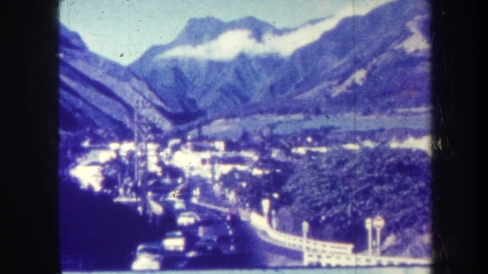Blue-cast 8mm view of a Hawaiian valley with roads, cars, small buildings, greenery, mountains, and cloud cover.
