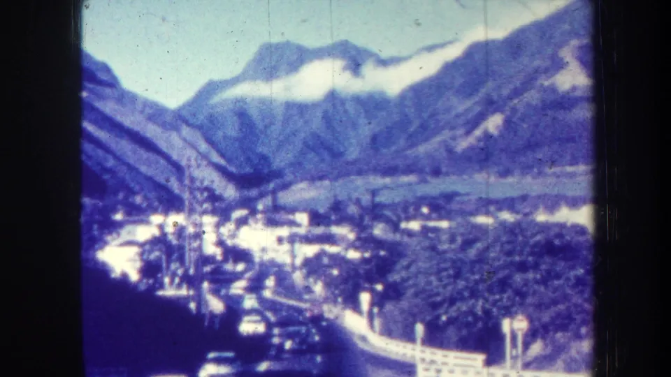 1967 Hawaii valley scene with cars on a curving road, white roadside railing, low buildings, lush vegetation, and mountains.