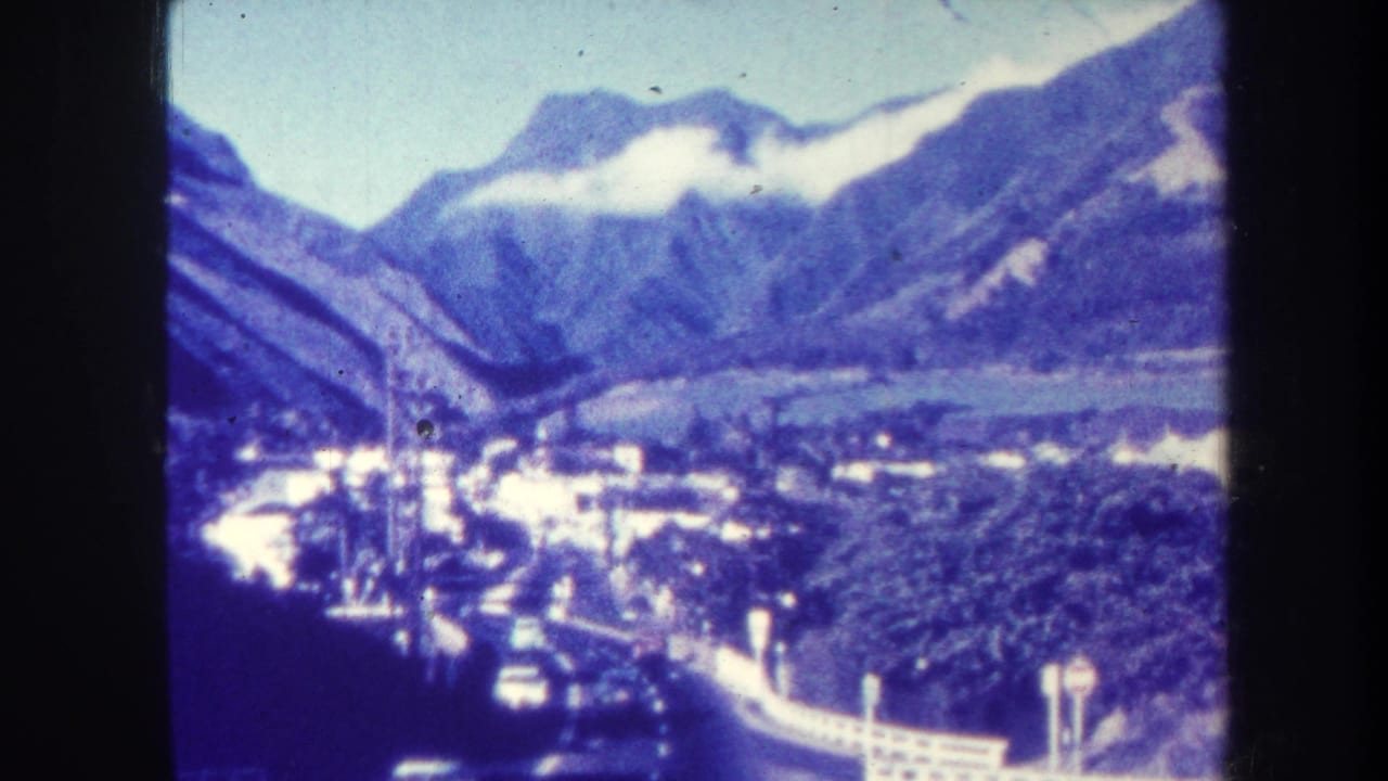 Mountain valley with town buildings, winding road, trees, and dark film-frame borders.