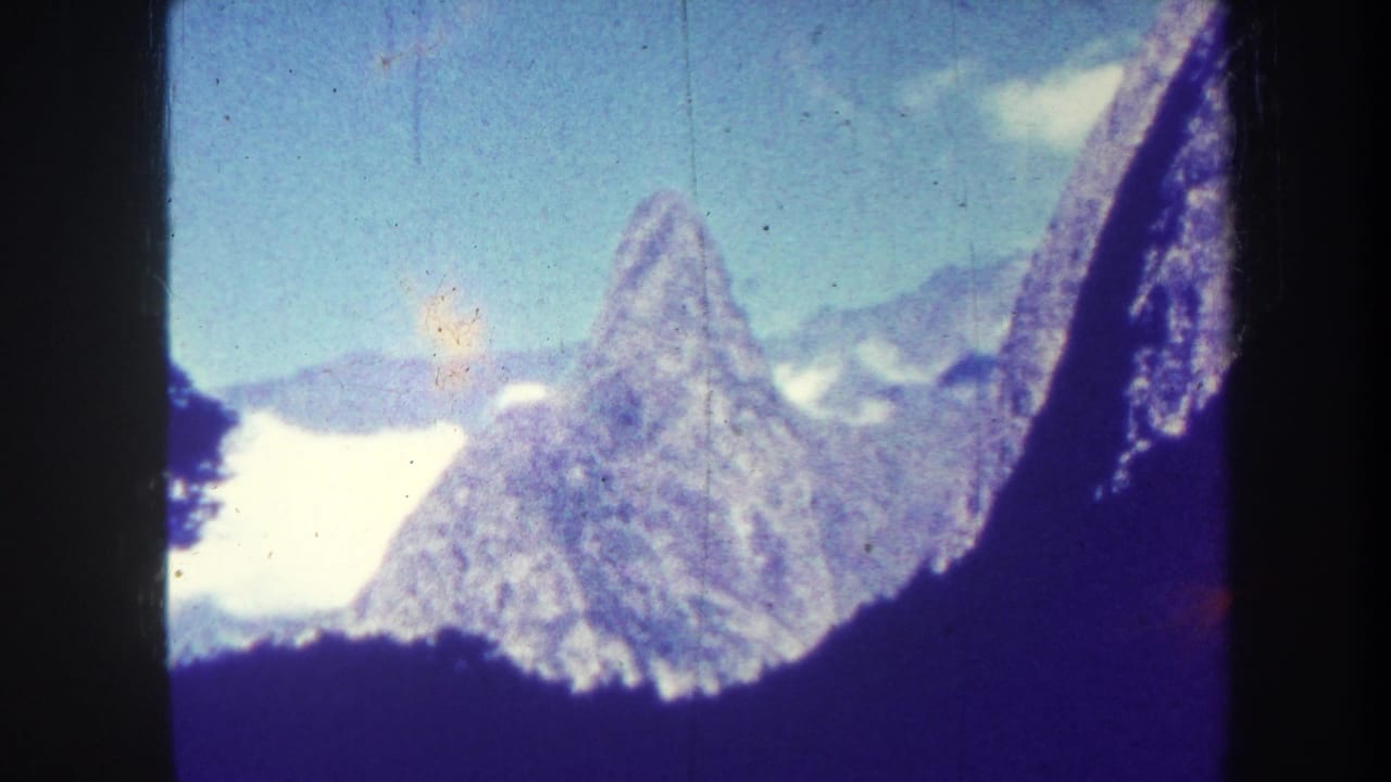 1967 8mm home-movie frame showing a distant jagged mountain peak, pale snow patches, blue sky, and dark foreground slopes.