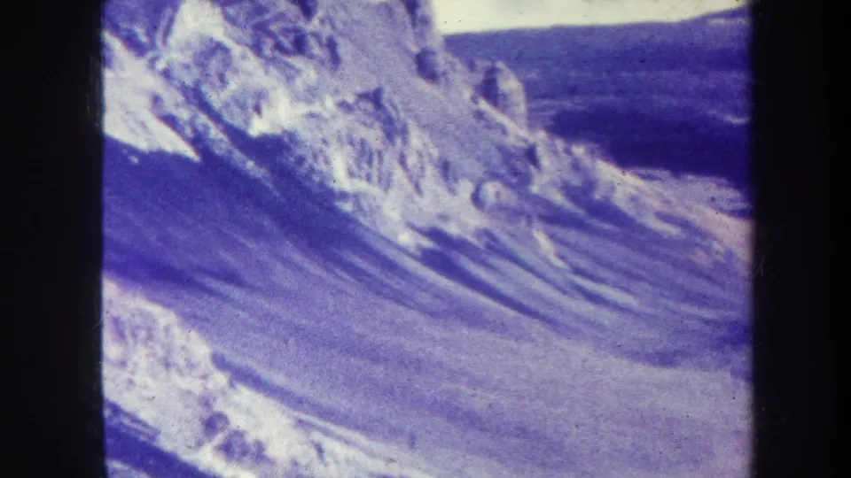 1967 8mm home-movie frame with a broad snow-streaked slope, rugged distant peak, blue sky, and visible scan borders.