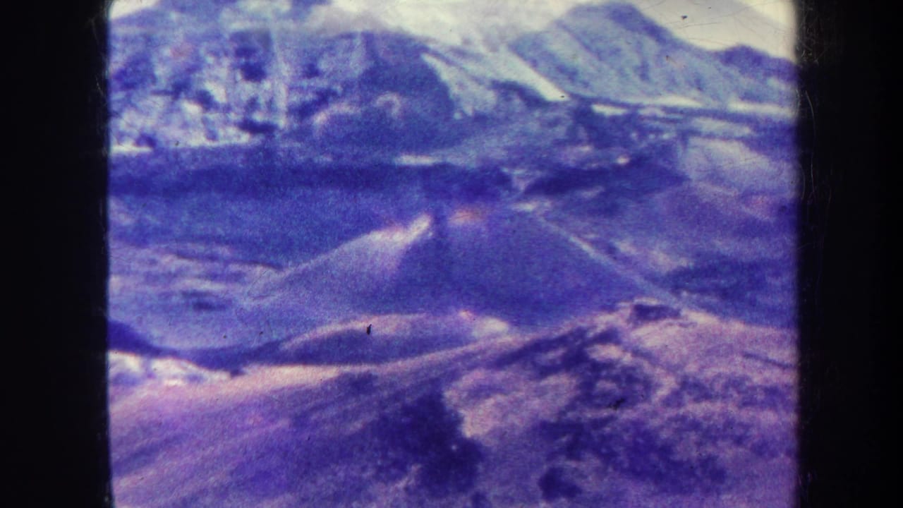 1967 8mm home-movie frame, metadata-labeled Honolulu, showing an aerial view of distant mountain ridges with snow-bright peaks and purple-blue film shift.