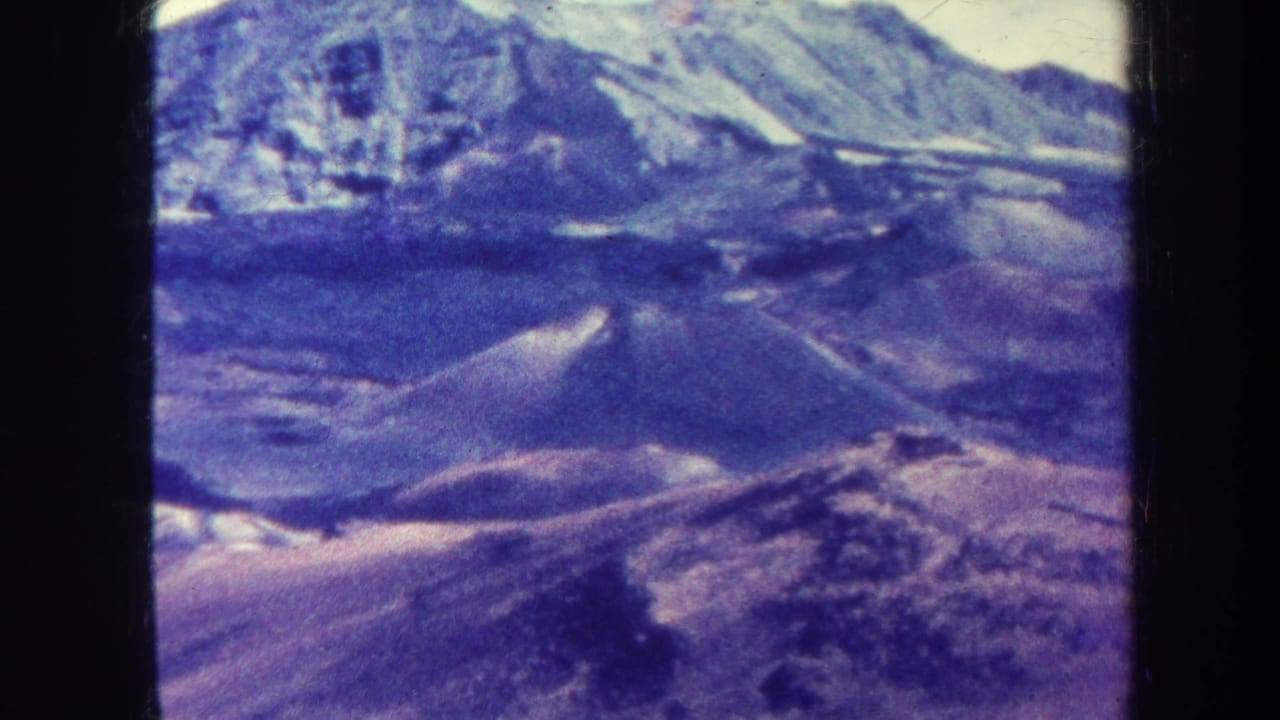 Aerial mountain landscape with dark ridges, pale sky, and visible 8mm frame edges.