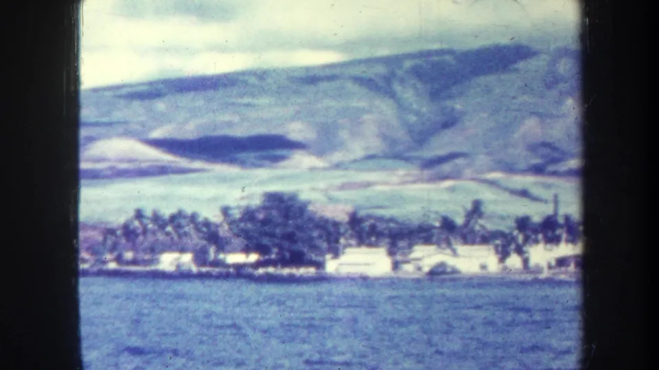 1967 Honolulu-area 8mm view across blue water toward low shoreline buildings, palm trees, and mountains beneath cloud cover.