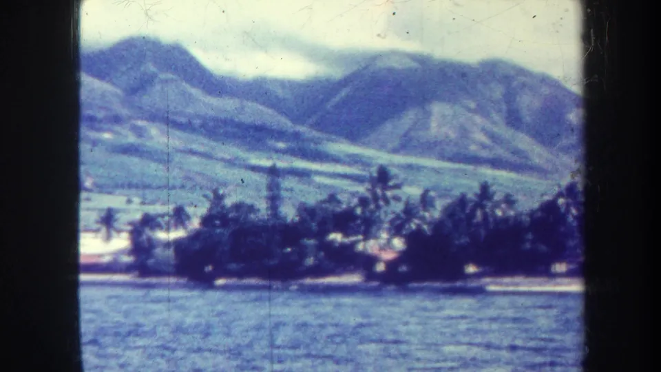 1967 Honolulu home-movie frame showing water, a darker palm-lined shore, low structures, and clouded mountains beyond.