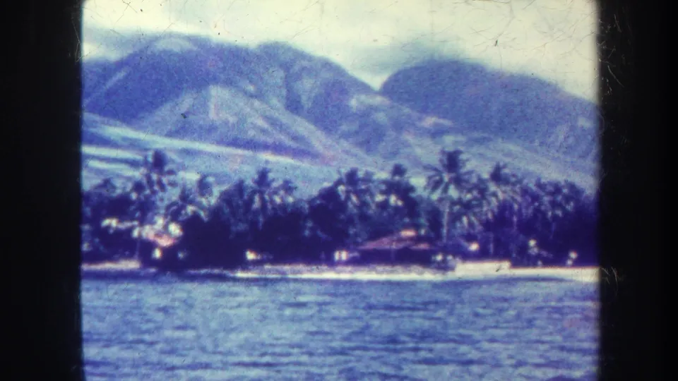 Soft 8mm frame from Honolulu in 1967 showing ocean water, a narrow beach, dense palms and mountain ridges.