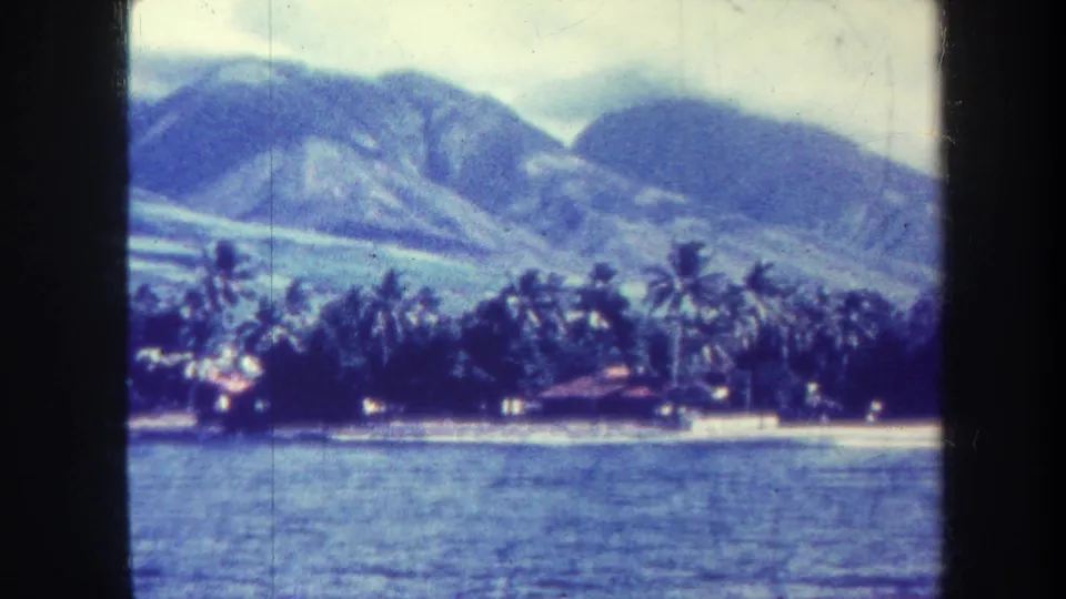 1967 Honolulu travel footage frame with a palm-covered shoreline, small waterfront structures and blue mountains beyond.