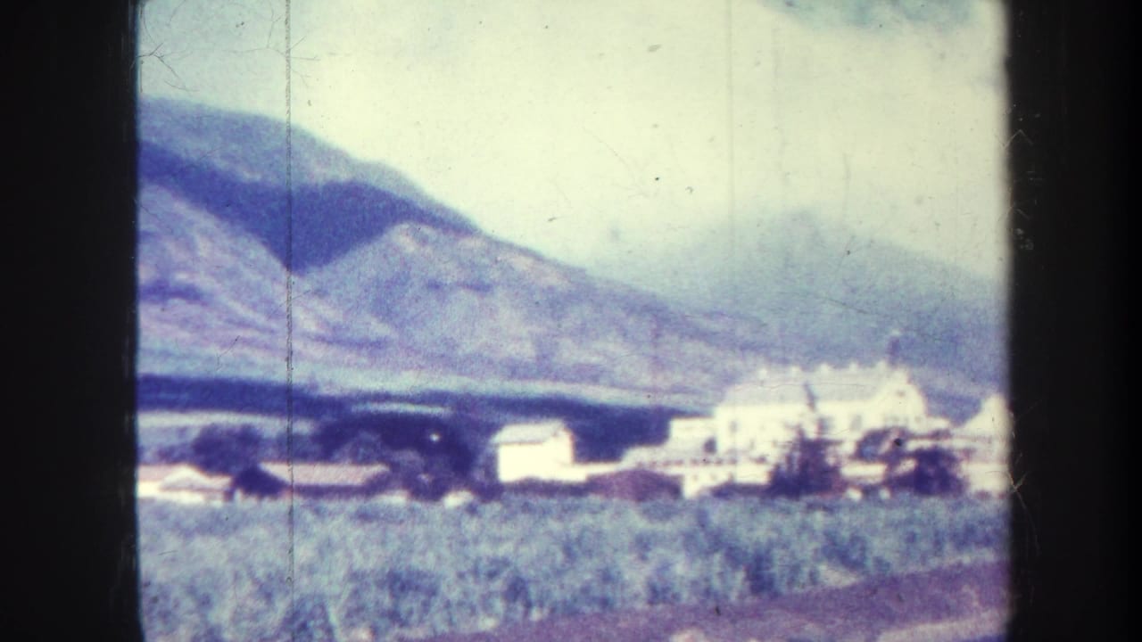 Distant industrial buildings and a smokestack below mountains, seen through an 8mm film frame.