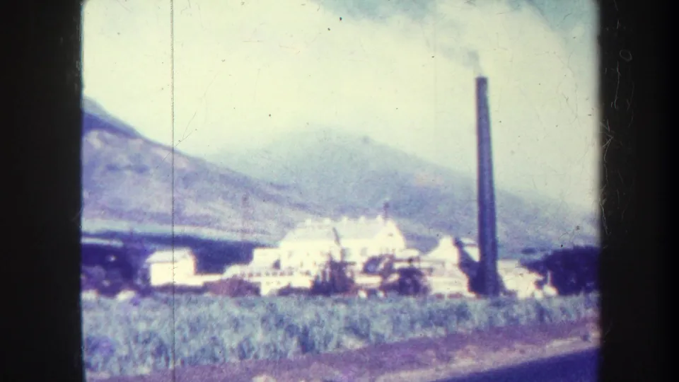 1967 Honolulu 8mm still showing an industrial site with smokestack, fields in front, and misty mountains behind.