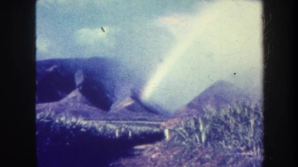 1967 Honolulu-area 8mm frame showing hazy mountain ridges, cloudy sky, dark foreground vegetation, and a bright diagonal band of light.