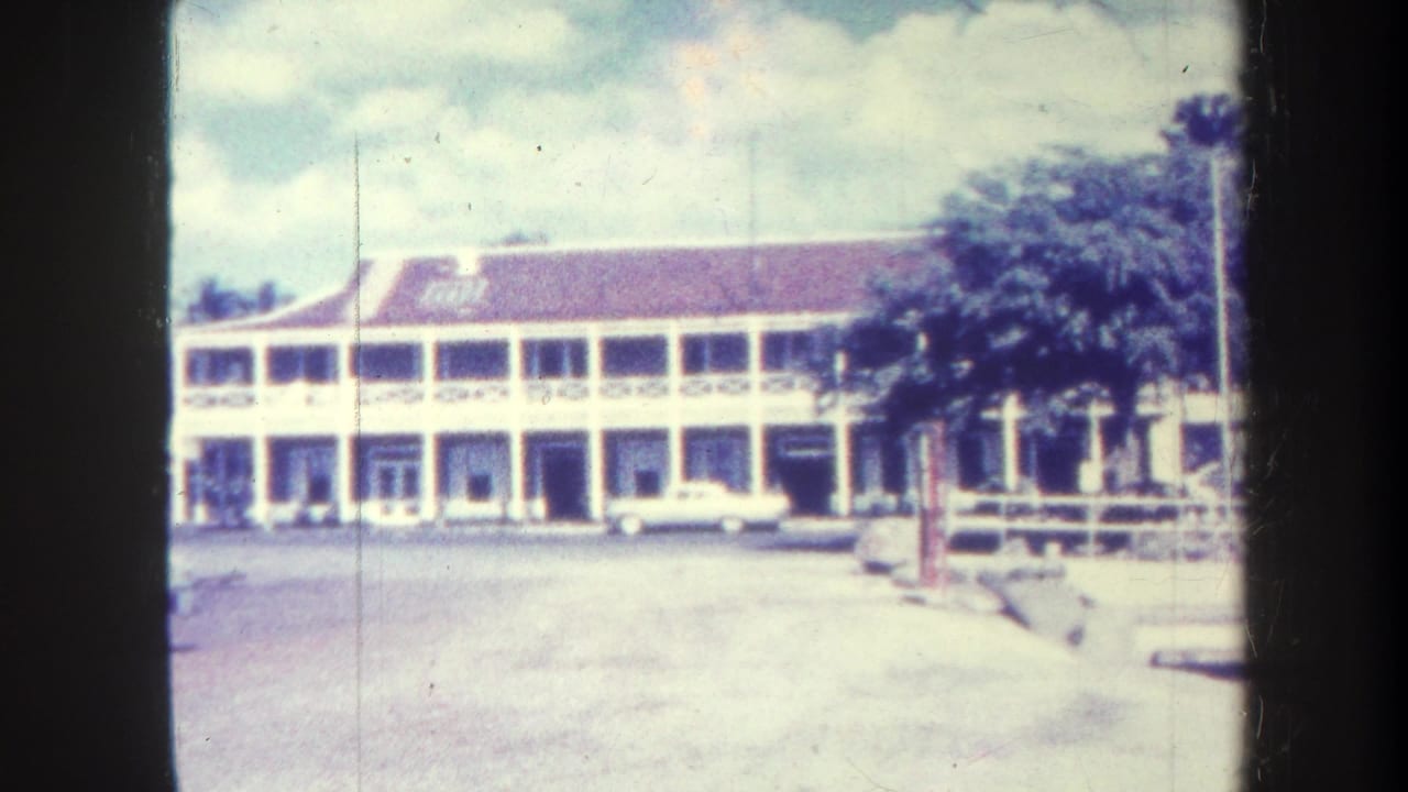 Two-story white building with a red roof, paved street, parked white car, and trees.