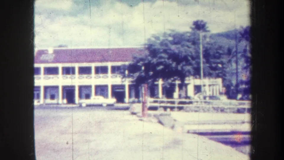 1967 Honolulu frame with the white two-story building, parked car, palm plantings, low wall, and mountain backdrop during the pan.