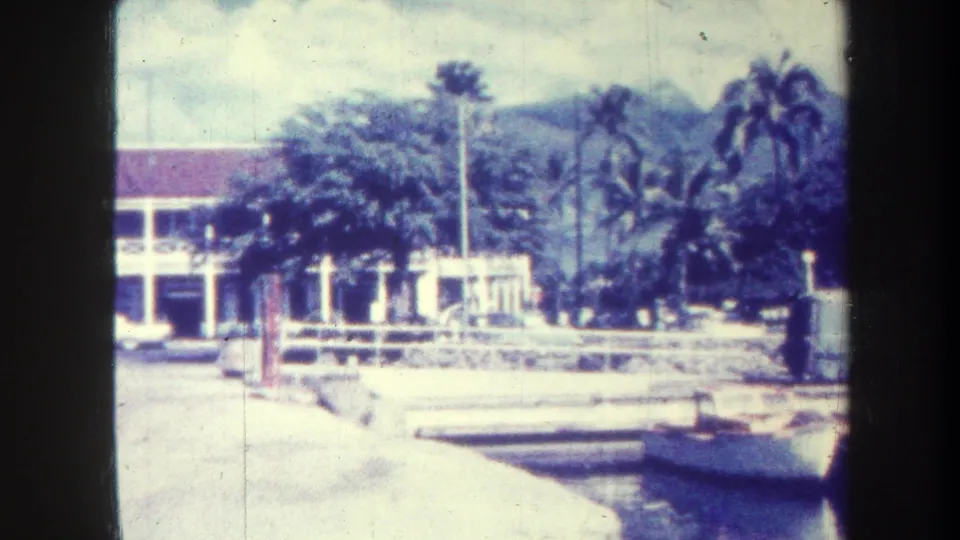 1967 Honolulu frame showing the pan near the building edge, with palm trees, low wall, paved street, distant mountains, and visible 8mm borders.