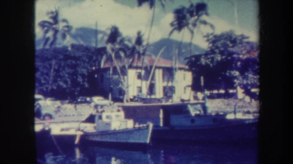 1967 Honolulu harbor view with small boats in the foreground, a pale waterfront building, palms, and green mountains beyond.