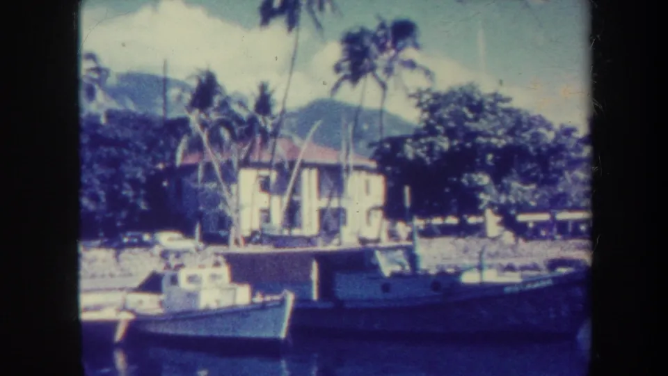Soft 8mm frame from Honolulu in 1967 showing moored small boats, palms, a waterfront building, and hazy mountain backdrop.