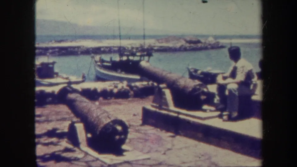 A man in light clothing sits beside weathered cannons on a Honolulu waterfront, with harbor boats and distant shoreline behind him.