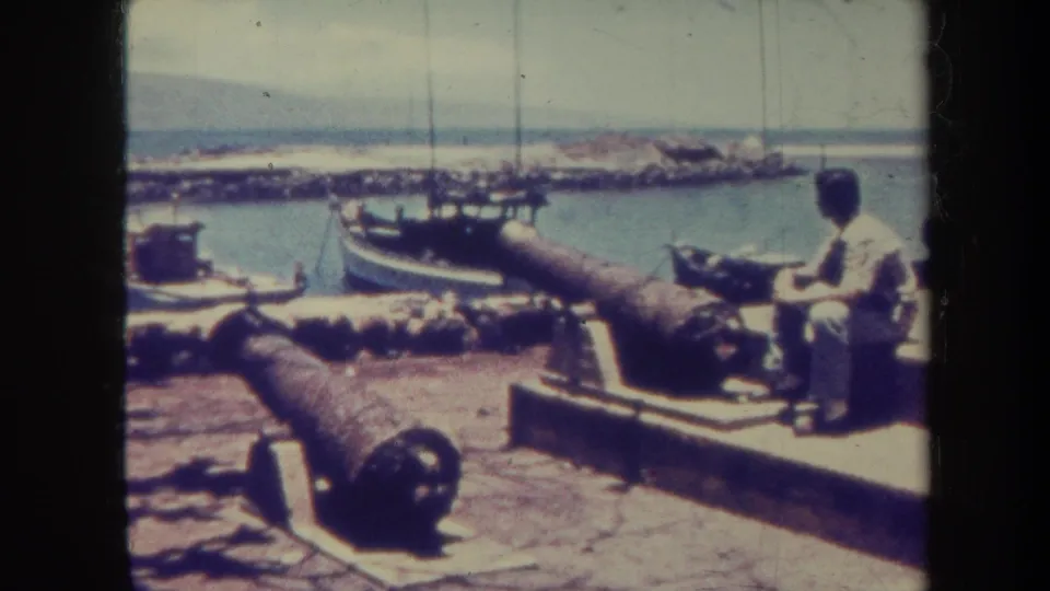 1967 Honolulu waterfront view with a seated man near two cannons, sailboats in the harbor, and distant mountains under hazy light.