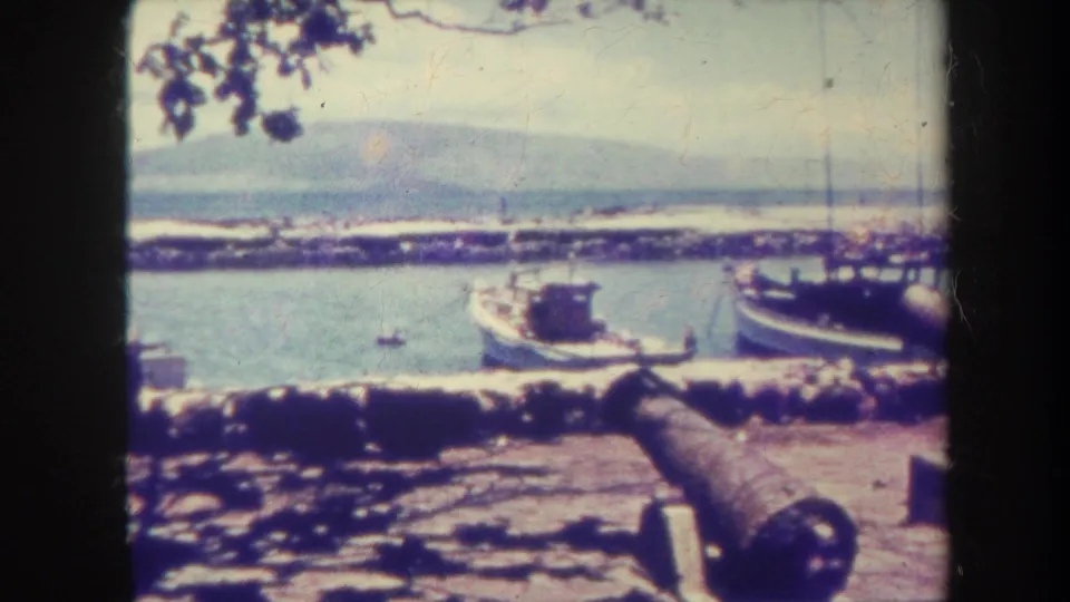 1967 Honolulu harbor footage showing an old cannon, docked boats, calm water, a stone breakwater, and distant hills.