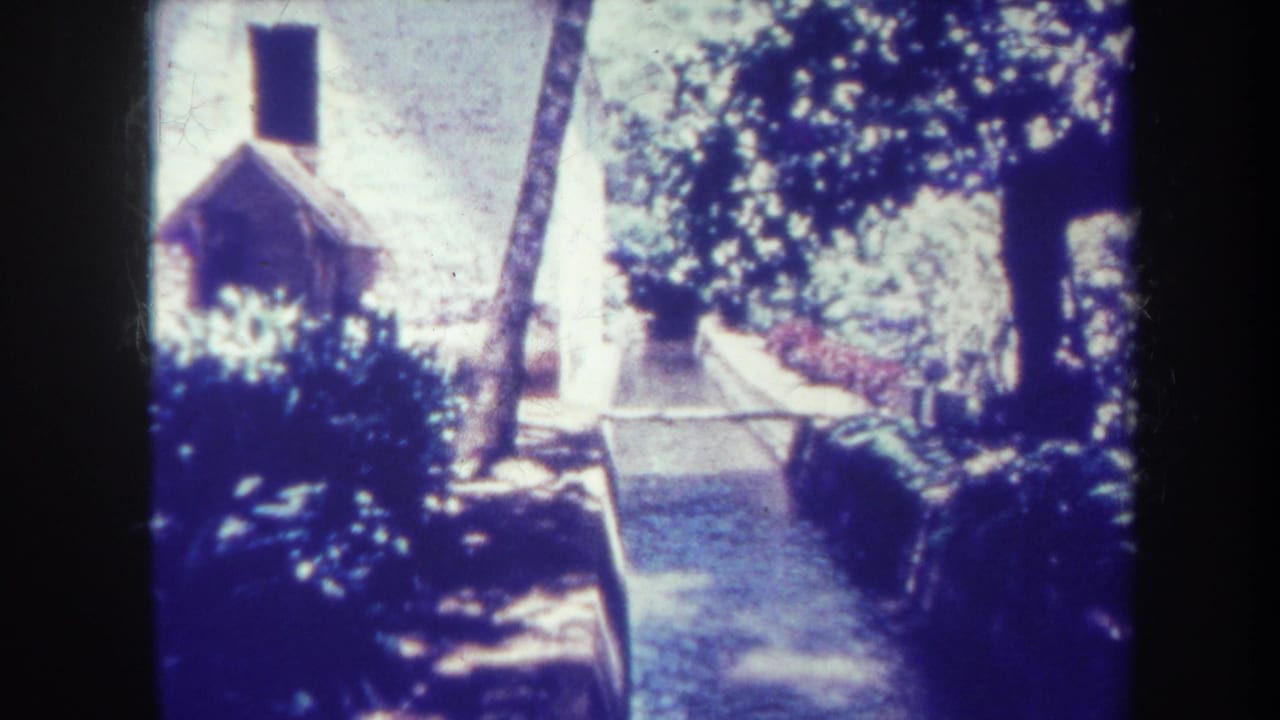 Small gabled building beside a paved path, shrubs, trees, and a low wall.
