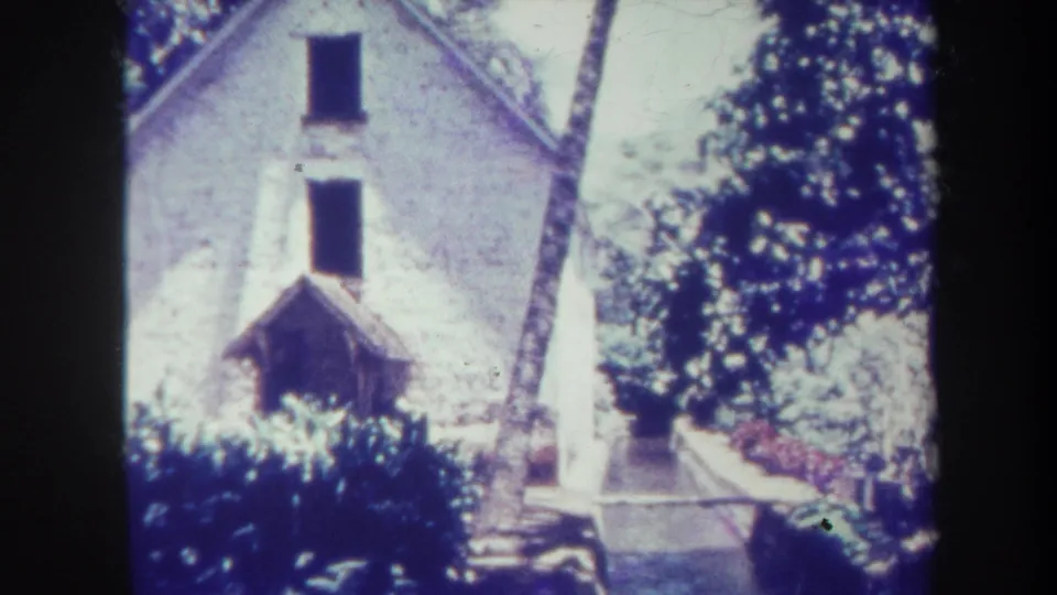 1967 Honolulu home-movie frame showing a pale gabled building with two dark windows, trees, shrubs, a path, and low stone edging.