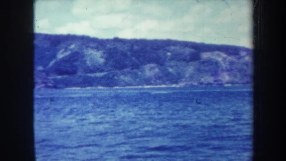 1967 Honolulu 8mm frame looking across blue water toward a low rugged mountain ridge under a pale sky.