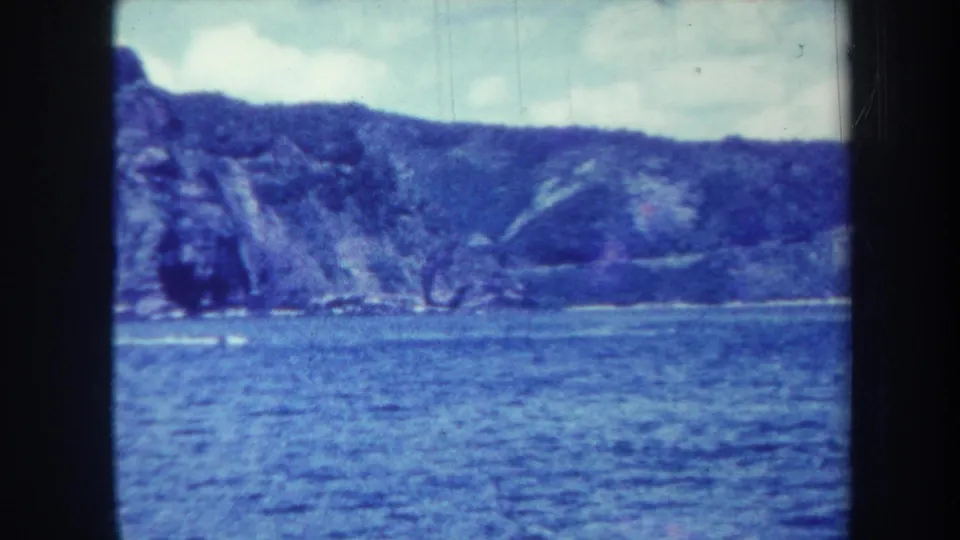 1967 Honolulu coastal frame with textured blue water, rugged mountain slopes, and soft cloudy sky in 8mm grain.