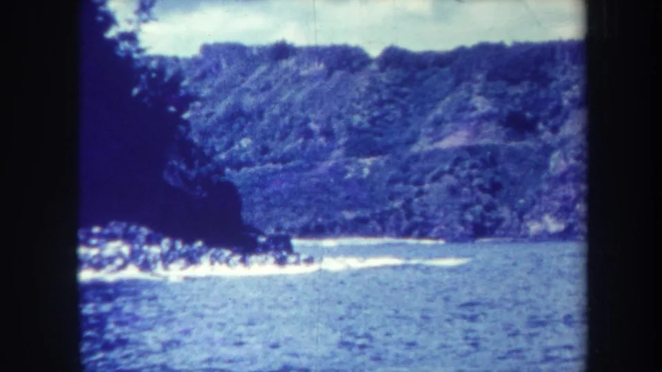 1967 Honolulu-area home-movie frame with a dark foreground edge, blue water, and a tree-covered cliff beyond.