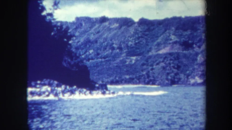1967 Honolulu home-movie frame showing a blue-tinted coastal cove, wooded cliff, water, and heavy 8mm frame edges.