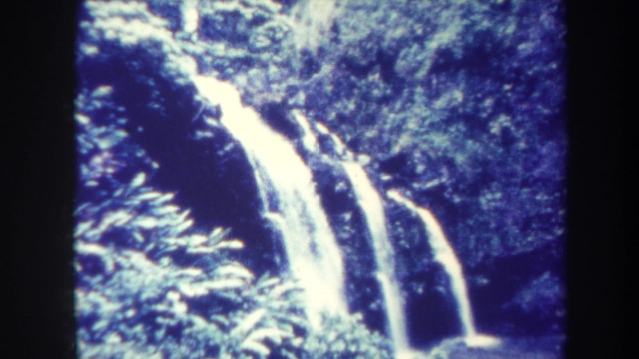 1967 Honolulu home-movie frame of a bright tropical waterfall dropping through dense forest vegetation into a shaded pool.